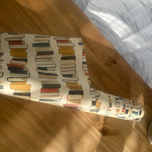 May include: A roll of white wrapping paper with a repeating pattern of colorful stacks of books. The paper is rolled up and lying on a wooden surface.