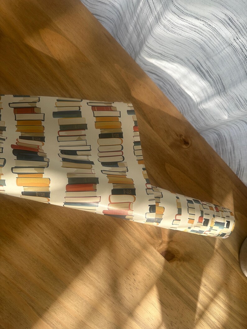 May include: A roll of white wrapping paper with a repeating pattern of colorful stacks of books. The paper is rolled up and lying on a wooden surface.