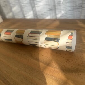 May include: A roll of wrapping paper with a pattern of stacked books in various colors and sizes. The paper is cream colored with brown, orange, and blue accents.