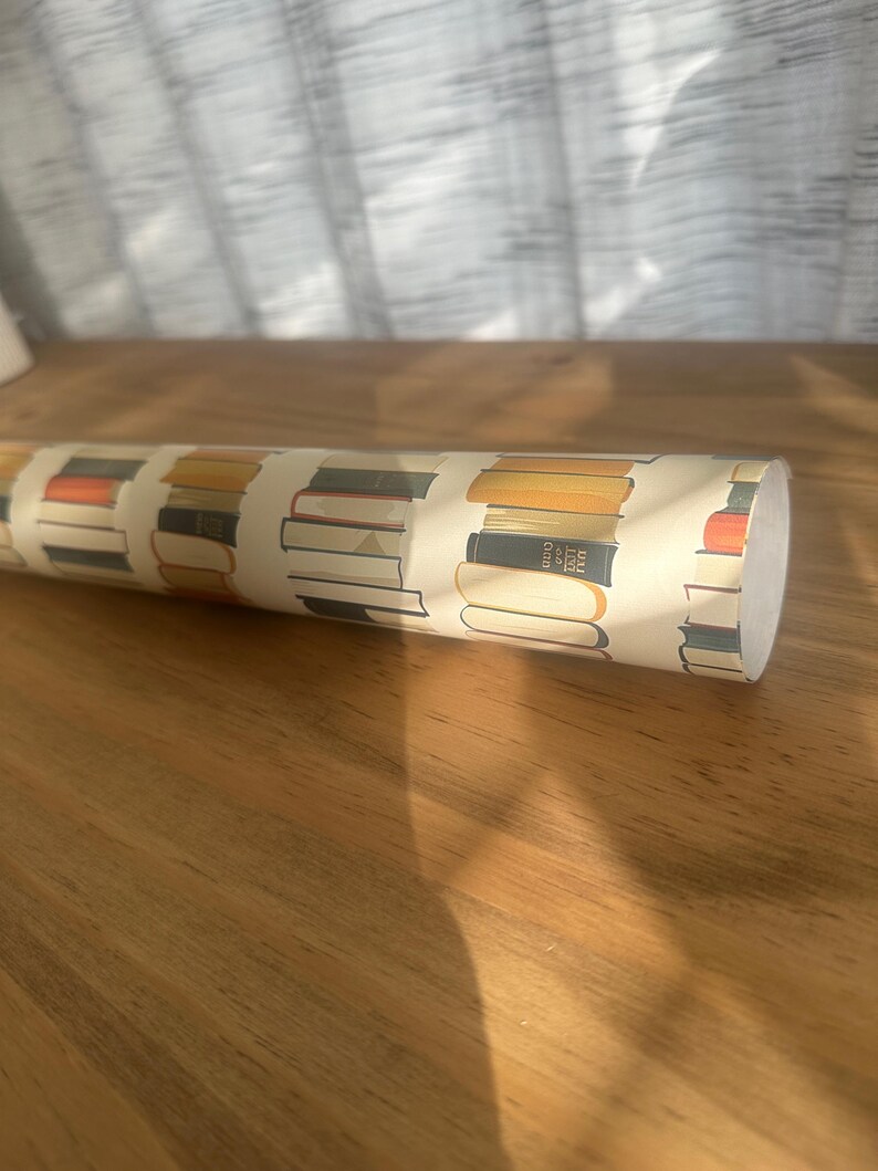 May include: A roll of wrapping paper with a pattern of stacked books in various colors and sizes. The paper is cream colored with brown, orange, and blue accents.
