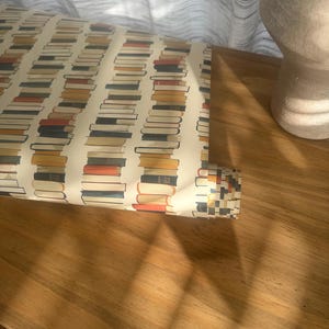 May include: A roll of wrapping paper with a pattern of stacked books in various colors, including red, blue, yellow, and brown. The paper is rolled up and partially unrolled, revealing the pattern.