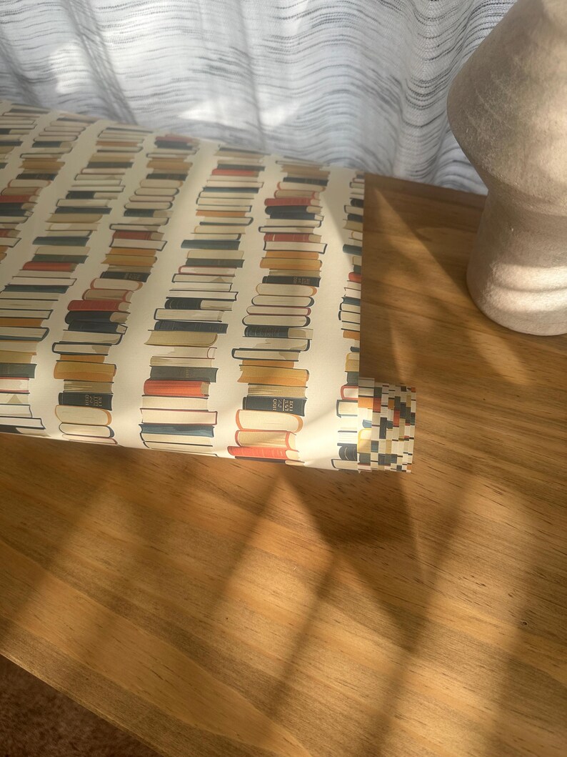 May include: A roll of wrapping paper with a pattern of stacked books in various colors, including red, blue, yellow, and brown. The paper is rolled up and partially unrolled, revealing the pattern.