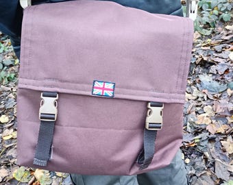May include: A brown messenger bag featuring a fold-over flap secured by two beige plastic side-release buckles with black nylon straps. A small rectangular patch displaying the Union Jack flag is centred on the front flap of the bag.