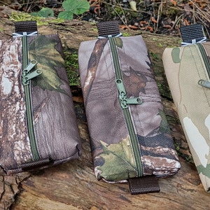 May include: Four camouflage-patterned zippered pouches with black straps. The pouches are lined up in a row on a brown wooden surface.
