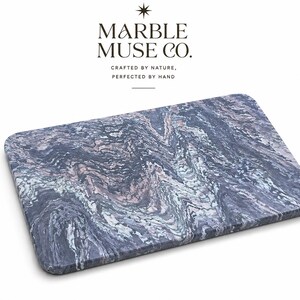 May include: A rectangular marble cutting board with rounded corners. The board features a swirling pattern of gray, blue, and pink hues. The text "MARBLE MUSE CO." is at the top, with the tagline "CRAFTED BY NATURE, PERFECTED BY HAND" below.