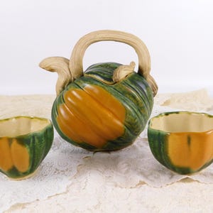 Puede incluir: Una tetera de cerámica y dos tazas a juego. La tetera y las tazas están pintadas con una combinación de colores verde y naranja. La tetera tiene un asa curva y una tapa. Las tazas tienen forma redondeada.