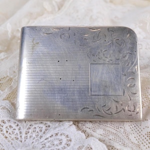 Może przedstawiać: Prostokątne, srebrzyste metalowe etui z zakrzywioną krawędzią. Etui posiada wytrawione wzory liniowe i kwiatowe. Tło to biała, koronkowa tkanina.