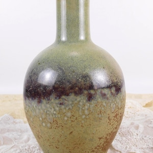 May include: A ceramic vase with a rounded body and a narrow neck. The vase features a gradient of colors, transitioning from a light green at the top to a dark brown band, and then to a speckled, earthy green at the base. The vase has a glossy finish.