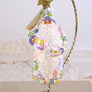 May include: Ornament of a Santa Claus figure, holding a teapot and teacup. The figure is adorned with a white beard and a floral pattern in shades of pink, purple, green, and yellow. A gold star tops the figure.