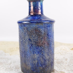 Puede incluir: Una botella de cerámica azul texturizada con cuello estrecho. La botella tiene un esmalte azul oscuro con reflejos azules claros y marrones. La superficie presenta un patrón vertical y acanalado. Podría ser un jarrón o un recipiente decorativo.