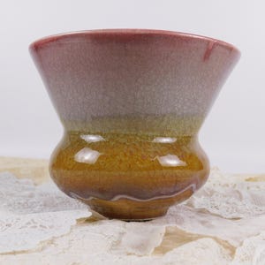 Może przedstawiać: Ręcznie wykonany ceramiczny wazon o unikalnym kształcie i błyszczącym wykończeniu. Wazon ma gradient kolorów, przechodzący od głębokiego czerwonobrązu na krawędzi do złocistobrązowej podstawy. Wazon stoi na białej koronkowej serwetce.