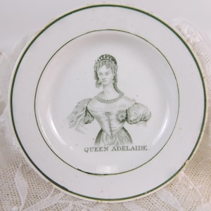 以下が含まれることがあります： 緑色の縁取りとアデレード女王の白黒肖像画が描かれた白いセラミックプレート。画像の下には「QUEEN ADELAIDE」の文字。白いレースの布の上に置かれています。