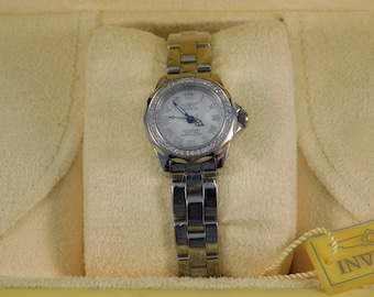 Puede incluir: Un reloj Invicta plateado con una esfera blanca y detalles de diamantes alrededor del bisel. El reloj está sobre un cojín blanco.