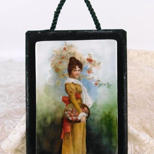 May include: A framed porcelain plaque featuring a woman in a yellow dress holding a basket of apples. The artwork has a black frame with a green rope handle. The woman is set against a backdrop of a tree with red apples.