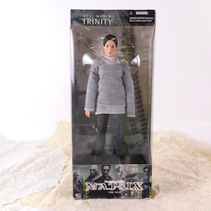 May include: A Trinity action figure from the Matrix film, packaged in a clear plastic box. The figure wears a gray sweater, black pants, and has short black hair. The box has the text "REAL WORLD TRINITY" and "MATRIX THE FILM".