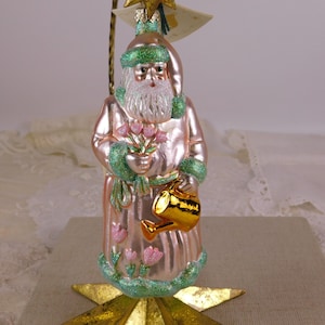 Può includere: Ornamento natalizio decorativo a forma di Babbo Natale. L'ornamento è rosa con accenti verdi e tiene in mano un mazzo di fiori rosa e un annaffiatoio dorato. L'ornamento è posto su una stella dorata.