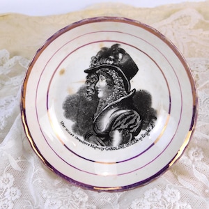 May include: Vintage ceramic plate featuring a portrait of a woman in a hat. The plate has a gold rim and a purple border with the text "Her most Gracious Majesty CAROLINE QUEEN of England".