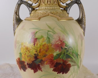 Op de afbeelding: Een grote, crèmekleurige porseleinen vaas met zilveren handvatten en gouden accenten. De vaas is versierd met een bloemmotief van roze, gele en rode bloemen.