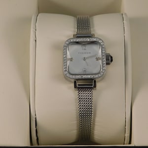 May include: A silver watch with a square face and a silver mesh band. The watch face has a white background with black Roman numeral hour markers and the word "TYCOON" printed in black. The watch is surrounded by a bezel set with clear crystals.