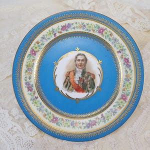May include: A decorative plate with a blue center featuring a portrait of a man in a military uniform. The plate has a floral border with gold trim. The outer rim is blue with gold detailing. A vintage collectible.