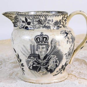 以下が含まれることがあります： クリーム色のアンティークピッチャー。王冠をかぶった人物の白黒ポートレートが描かれています。縁と側面に花と葉の模様があり、曲がった取っ手と注ぎ口が付いています。