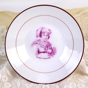 May include: A white ceramic plate featuring a portrait of Queen Caroline in purple tones, with a maroon rim and a decorative maroon inner circle. The text "Queen Caroline" is visible below the portrait.