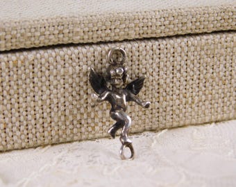 Puede incluir: Un pequeño dije de ángel plateado con los brazos extendidos y alas detalladas. El dije tiene un lazo en la parte superior para sujetarlo y una estética vintage delicada. El fondo presenta una superficie beige texturizada y encaje blanco.