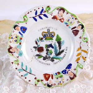 以下が含まれることがあります： 白いセラミックプレート、縁はスカラップ模様で、手描きのディテールが施されています。「CORONATION AT WESTMINSTER SEPTEMBER 8 1831」と書かれた王冠のエンブレムが中央にあります。縁には花柄や人物の装飾が施されています。