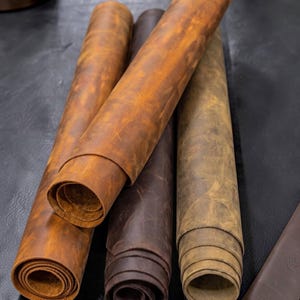 Peut inclure: Quatre rouleaux de cuir dans des nuances variées de brun, incluant le beige clair, le brun moyen et le chocolat foncé. Le cuir présente une finition mate et vieillie avec des motifs de grain et des textures naturels visibles.