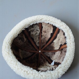 May include: A winter hat with a white, fluffy, sheepskin-like brim and a brown and beige patterned interior. The hat's interior features brown leather-like strips radiating from a central point.