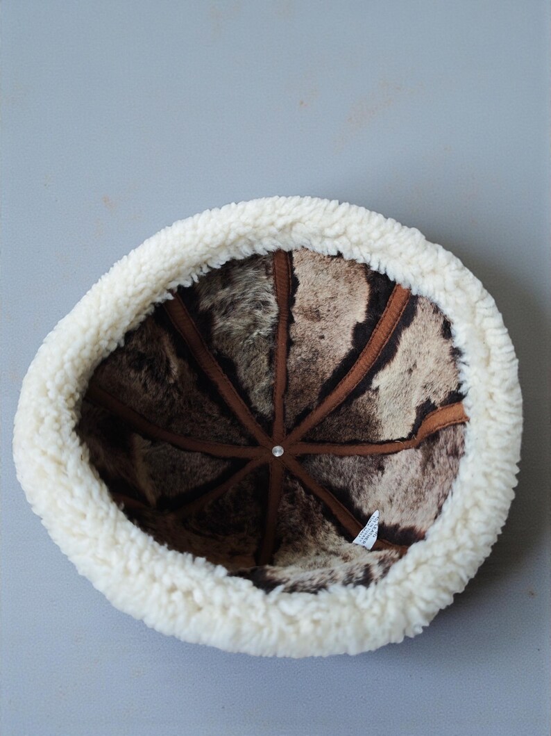 May include: A winter hat with a white, fluffy, sheepskin-like brim and a brown and beige patterned interior. The hat's interior features brown leather-like strips radiating from a central point.