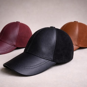 May include: Three leather baseball caps in burgundy, black, and brown. The caps have a classic six-panel design with a curved brim. The black cap has a suede panel. The caps are displayed on a neutral background.