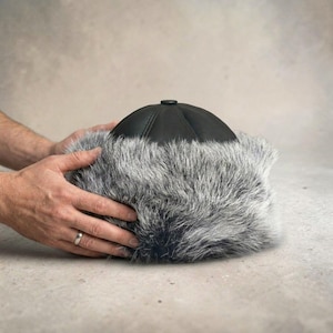 May include: A black leather cap featuring a thick, textured band of grey and black faux fur around the base. The crown of the hat is constructed from smooth, dark leather panels that meet at a central button detail on the top of the headwear.