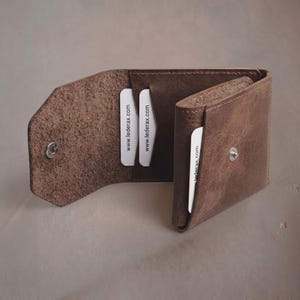 May include: A brown leather wallet with a textured surface, featuring a silver metal snap button closure. The interior contains multiple card slots, each holding a white card printed with the text www.lederax.com in black lettering.
