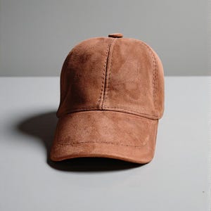 May include: A brown suede baseball cap with a curved peak. The cap features a classic six-panel design, a top button, and stitched seams. The hat is a solid colour and appears to be made of soft material.