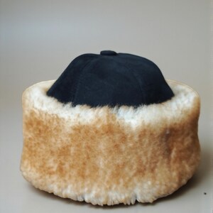 May include: A black hat with a rounded crown and a fluffy, tan-colored fur trim. The hat appears to be made of a soft material and is designed to keep the wearer warm. The fur trim is thick and plush.