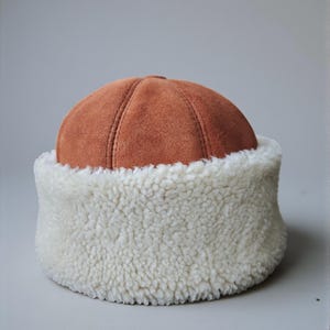 May include: A brown suede hat with a white, fluffy, shearling-style brim. The hat has a rounded crown with vertical seams. The brim is thick and wraps around the entire base of the hat, creating a warm and cozy look.