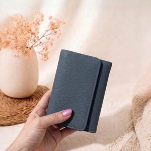 Pode incluir: Uma carteira de couro azul ardósia com uma superfície texturizada. A carteira é segurada por uma mão. Um vaso com flores secas está ao fundo. A carteira tem cerca de 10 cm de altura.