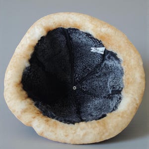 May include: A winter hat with a light brown exterior and a dark gray interior. The hat has a soft, furry texture and a circular shape. The brim is a lighter shade of brown, and the interior appears to be lined with a dark, plush material.