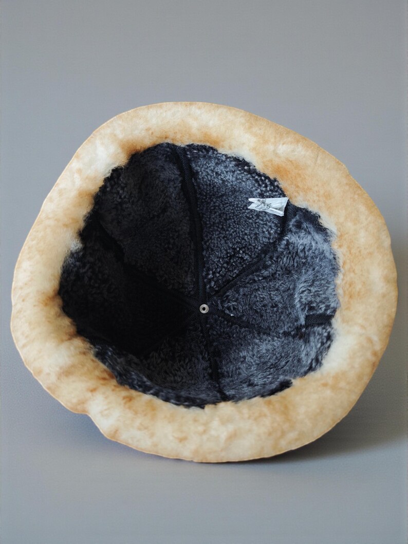 May include: A winter hat with a light brown exterior and a dark gray interior. The hat has a soft, furry texture and a circular shape. The brim is a lighter shade of brown, and the interior appears to be lined with a dark, plush material.