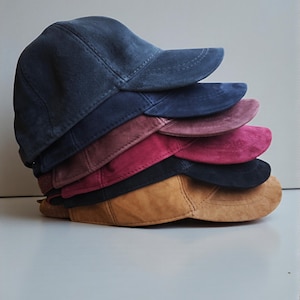 Könnte beinhalten: Ein Stapel von sechs Baseballkappen in verschiedenen Farben, darunter Marineblau, Burgunderrot und Beige. Die Kappen bestehen aus einem weichen, Wildleder-ähnlichen Material und haben einen klassischen, gebogenen Schirm. Die Kappen sind aufeinander gestapelt.
