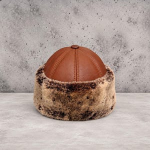 May include: A round hat featuring a smooth, brown leather crown with visible stitched seams and a wide, thick band of textured, mottled brown faux fur encircling the base. The design is structured with a small button at the very top centre.