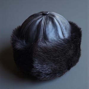 Puede incluir: Un sombrero negro con la parte superior redondeada y un ribete de piel. La copa del sombrero está hecha de un material oscuro y liso con costuras verticales. El ribete de piel es un material oscuro y texturizado que rodea la base del sombrero.