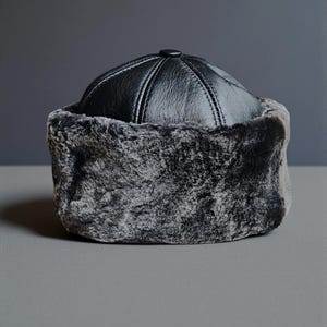 May include: A black leather hat with a fur trim. The hat has a rounded top with stitched panels and a small button on top. The fur trim is a dark grey colour. The hat is on a grey surface.