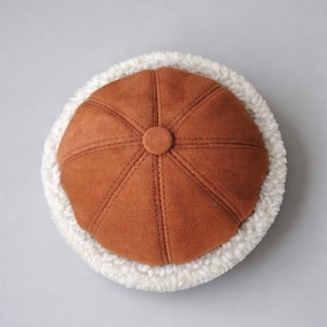 May include: A brown suede-like hat with a button on top and stitched details. The hat is surrounded by a fluffy, white, faux fur trim. The hat is a winter accessory.