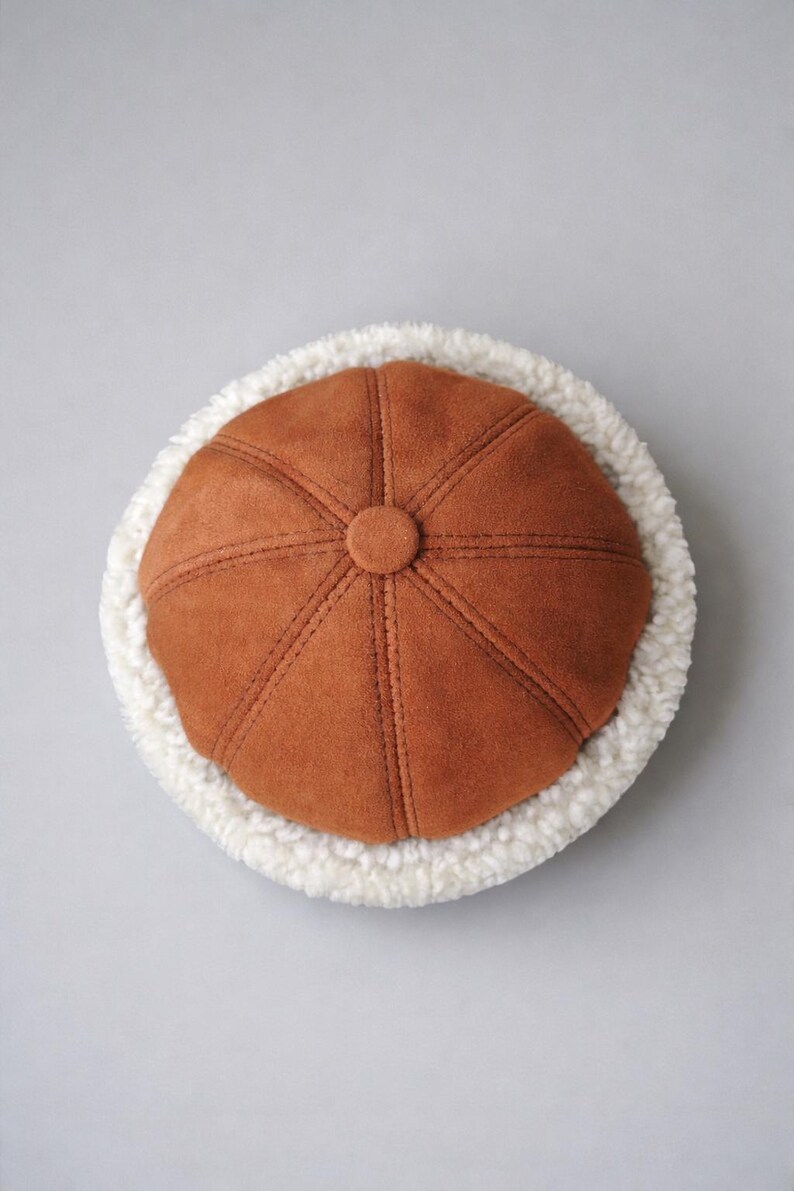 May include: A brown suede-like hat with a button on top and stitched details. The hat is surrounded by a fluffy, white, faux fur trim. The hat is a winter accessory.