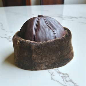 Op de afbeelding: Een bruine leren hoed met een bontrand. De hoed heeft een afgeronde kroon met een knoop bovenop en een gestikte naad in het midden. De bontrand is een donkerdere bruintint. De hoed is waarschijnlijk ontworpen voor koud weer.