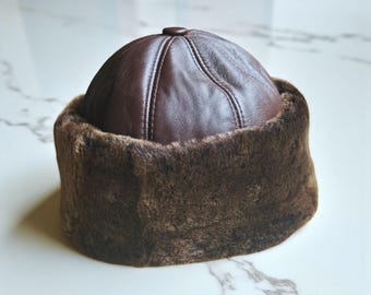 May include: A brown leather cap featuring a rounded crown with stitched paneling and a central button detail. The base of the cap is encircled by a thick, textured brown faux fur band that provides a contrasting finish to the smooth leather.