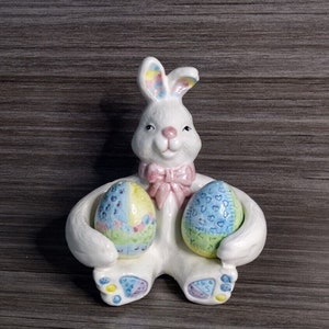 May include: A white ceramic Easter bunny figurine holding two colorful Easter eggs. The bunny has pink ears and a pink bow. The eggs are decorated with blue, green, yellow, and pink patterns.