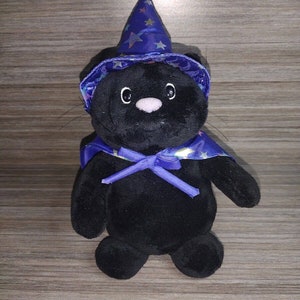 Könnte beinhalten: Schwarze Plüschkatze mit einem lila und blauen Zaubererhut mit Sternen. Die Katze sitzt auf einer Holzoberfläche.
